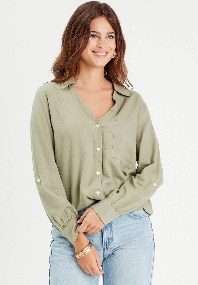 Blouse vert clair avec des manches longues, fermeture à boutons et une poche. Fabriquée en tissu doux, elle présente une coupe décontractée et des poignets retroussables.