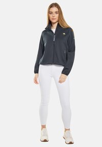 Svart zip-up jacka med hög krage, med en grön logotyp på bröstet och en sidoficka. Bärs med vita leggings och sneakers.