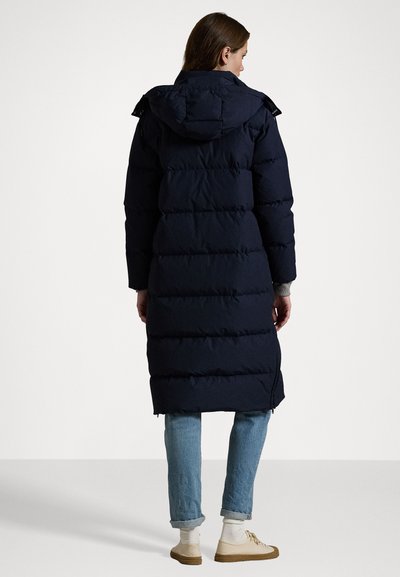 Geacă puffer bleumarin cu glugă, cu un design matlasat, de lungime medie, și buzunare laterale cu fermoar. Purtați-o peste blugi și adidași de culoare deschisă.