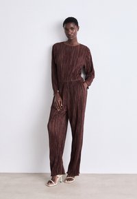 Combinaison bourgogne avec des rayures verticales texturées, manches longues, taille cintrée, jambes larges et fentes à l'ourlet. Portée avec des sandales à talons hauts beiges.