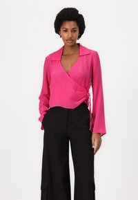 Pink wrap blouse with long sleeves and a notched collar, featuring a textured fabric and side tie detail, paired with high-waisted black trousers.