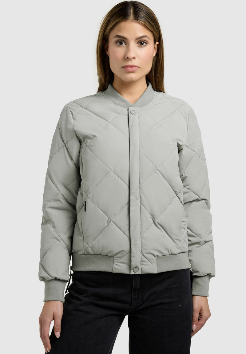Femme portant une veste bomber matelassée gris clair avec des poignets et un ourlet côtelés, associée à un pantalon noir, debout devant un fond uni.