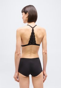 Γυναίκα με κοντά μαλλιά, φορώντας μαύρο δαντελωτό bralette με racerback και μαύρο σορτς, στεκόμενη με την πλάτη προς τα έξω μπροστά σε απλό φόντο.