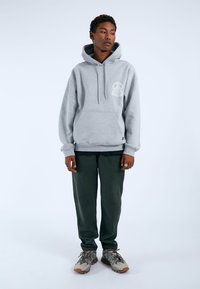 Grå huvtröja i mjukt material, med en framficka och dragsnodd. Kombinerad med mörkgröna byxor och flerfärgade sneakers.