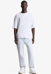 Mladic stoji, odjeven u običnu bijelu oversized majicu, svijetloplave traperice, bijele tenisice i tanku ogrlicu, nasuprot bijeloj pozadini.