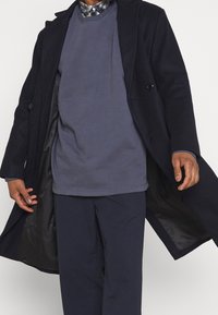 Abrigo largo azul marino con un exterior texturizado similar a la lana, diseño de doble botonadura y detalles de botones, llevado sobre un sudadera de cuello redondo azul marino y pantalones.