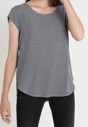 Vrouw draagt een zwart-wit gebloemde blouse met korte mouwen en ronde hals, zwarte broek, arm geheven bij het hoofd.