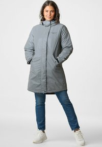Grijze lange parka jas met een rits aan de voorkant, capuchon en zijzakken. Heeft een zachte textuur en eenvoudig ontwerp, gedragen over blauwe spijkerbroeken en witte sneakers.