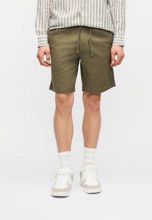 Άνδρας που φοράει σορτς ελιάς με κορδόνι, λευκές κάλτσες, λευκά αθλητικά παπούτσια και γκρι-λευκό ριγέ πουκάμισο με μακριά μανίκια, στέκεται μπροστά σε απλό φόντο.