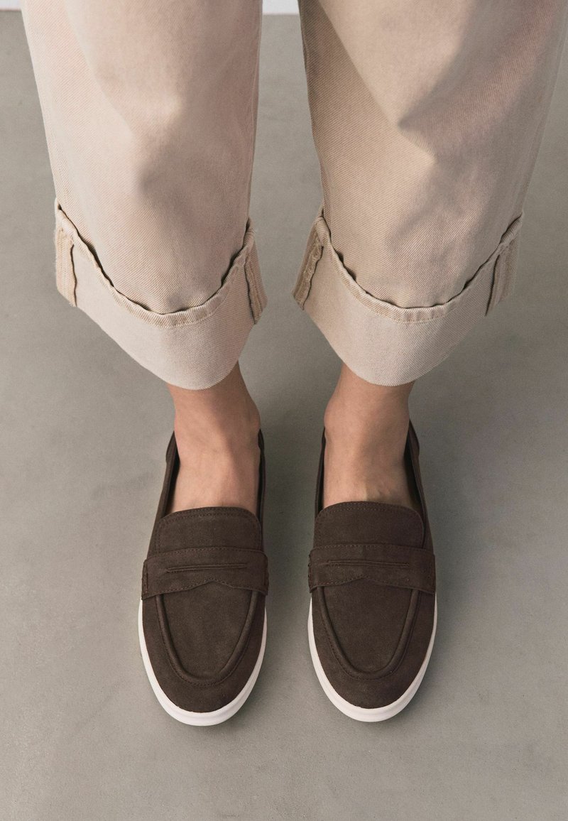 Jambes portant un pantalon beige retroussé avec des mocassins marron foncé sans lacets dotés de semelles blanches sur un sol gris.