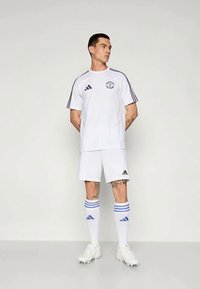 Biele futbalové uniformy adidas so značkou na tričku, modrými pruhmi na rukávoch, zodpovedajúcimi šortkami a pruhovanými ponožkami.