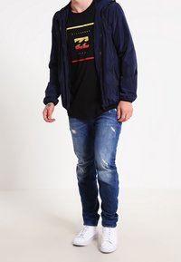 Marinblå vindjacka, svart T-shirt med färgglada grafikdetaljer, slitna blå jeans och vita sneakers. Enkel, avslappnad outfit.