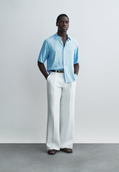 Νεαρός άνδρας που στέκεται φορώντας ανοιχτό μπλε πουκάμισο με σχέδιο και κοντά μανίκια, λευκό παντελόνι με φαρδιές παντελόνες, μαύρη ζώνη και μαύρα σανδάλια μπροστά σε απλό φόντο.