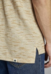 Camisa de textura beige claro con un patrón sutil, que presenta aberturas laterales y una pequeña etiqueta de logotipo, combinada con jeans de mezclilla azul.