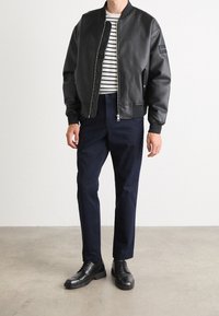 Svart bomberjacka i konstläder med ribbade ärmslut, tillsammans med en randig t-shirt och marinblå byxor. Svarta snörskor fulländar outfiten.