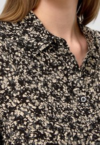 Camicia a maniche corte nera e beige con motivo, caratterizzata da un colletto, chiusura a bottoni e un tessuto testurizzato. I motivi consistono in forme astratte.