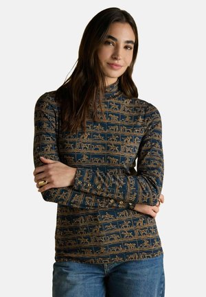 Frau mit langen, dunklen Haaren, die einen navyblauen und beigen gemusterten Rollkragenpullover mit langen Ärmeln und blaue Jeans trägt, mit gekreuzten Armen und sanftem Lächeln.