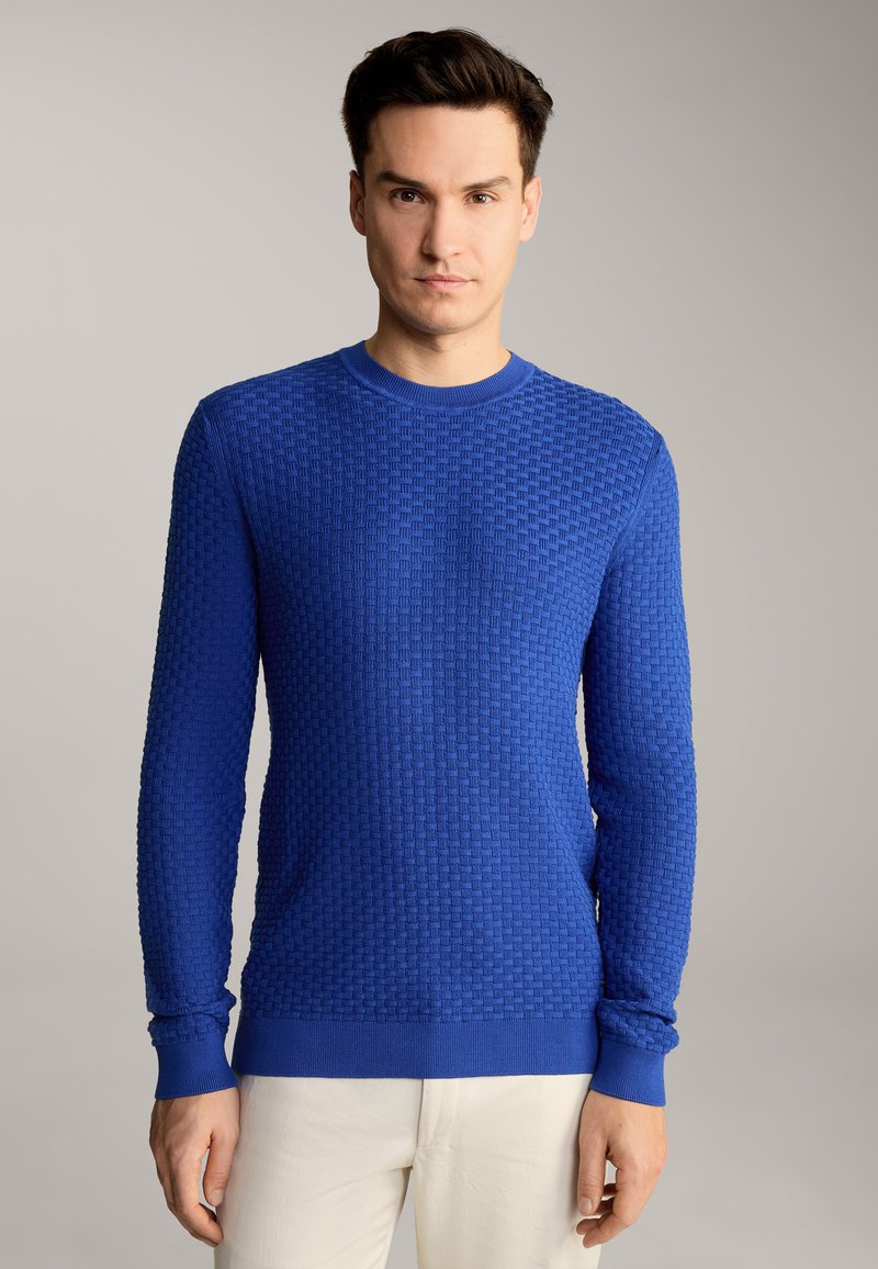 Pull en tricot bleu avec un motif texturé, encolure ronde et manches longues. Caractérisé par des poignets et un ourlet côtelés, associé à un pantalon de couleur claire.