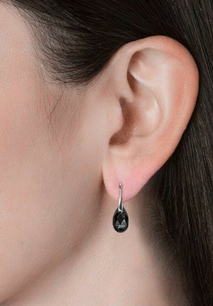 Orecchini a goccia nera con un design sfaccettato, sospesi da ganci in tonalità argentata, indossati all'orecchio di un modello su una pelle liscia.