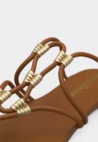 Καφέ επίπεδη σαγιονάρα με λεπτές λουρίδες τυλιγμένες σε χρυσές μεταλλικές ταινίες πάνω στο πόδι.