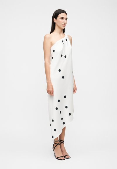 Γυναίκα που φοράει λευκό φόρεμα με ένα ώμο και μαύρες πουά, καθώς και μαύρα ψηλοτάκουνα σανδάλια με λουράκια, μπροστά σε απλό φόντο.