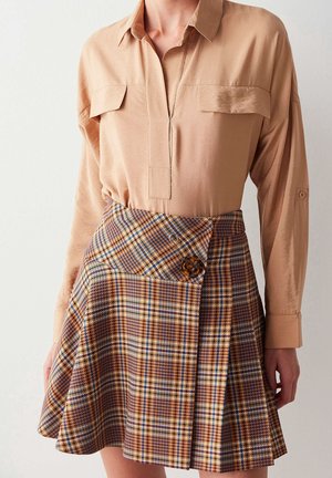 Femme portant une chemise beige à manches longues rentrée dans une jupe à carreaux taille haute avec un détail de bouton en écaille.