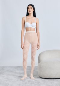 Καλσόν από δαντέλα σε γυμνό χρώμα με λουλουδένιο σχέδιο, ψηλοκάβαλο κόψιμο και ομαλή υφή, σε συνδυασμό με ένα λευκό στηθόδεσμο με ενίσχυση, πάνω σε ουδέτερο φόντο.