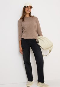 Femme portant une casquette et un haut beiges, un pantalon noir, tenant un grand sac beige, se tenant contre un mur blanc, souriante.