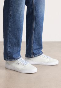 Personne portant un jean en denim bleu et des baskets blanches basses avec des lacets à motifs bleus, se tenant sur un sol beige.