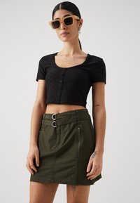 Svart cropped topp med knappe detaljer, sammen med en olivengrønn nederdel med belte i livet og glidelåslommer. Klare rektangulære solbriller.
