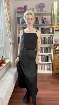Schwarzes, ärmelloses Maxikleid mit enganliegendem Oberteil und fließendem, strukturiertem Rock. Getragen mit klobigen schwarzen Stiefeln. Bücherregale im Hintergrund.