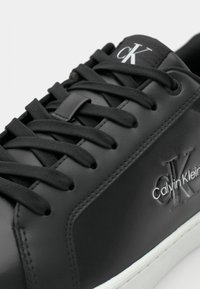 Zwarte leren sneakers met platte zwarte veters, een gestructureerd oppervlak en een witte rubberen zool. Voorzien van het zilveren logo "Calvin Klein" en "CK" accenten.