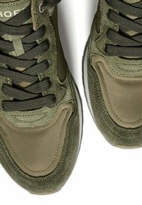 Sneaker verde oliva con una combinazione di materiali in suede e nylon, con lacci neri e dettagli texturizzati lungo i lati. Suola in gomma piatta.