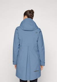 Blå vanntett jakke med hette, lange ermer og plissert design bak. Har en liten logo og kontrastdetalj nær kanten.