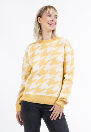 Sárga és fehér houndstooth mintázatú pulóver, kötött textúrával, bordázott nyakkivágással és mandzsettákkal, sötét farmerral párosítva.