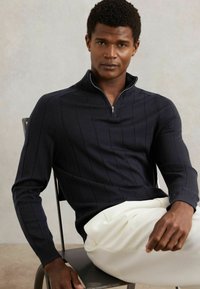 Granatowy sweter z dzianiny z pionowymi paskami, z kołnierzem na połowę zamka i prążkowaną fakturą; noszony z białymi spodniami, siedzący na krześle.