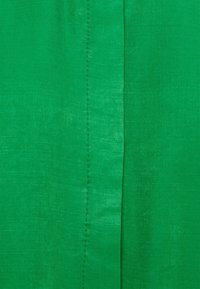 Tessuto verde con una superficie liscia, caratterizzato da un leggero luccichio. Una cucitura verticale corre lungo un lato, indicando i dettagli della sua costruzione.