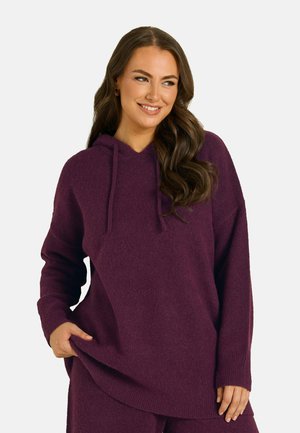 Femme portant un sweat à capuche violet ample à manches longues et un pantalon assorti, souriante et regardant sur le côté, une main dans la poche.