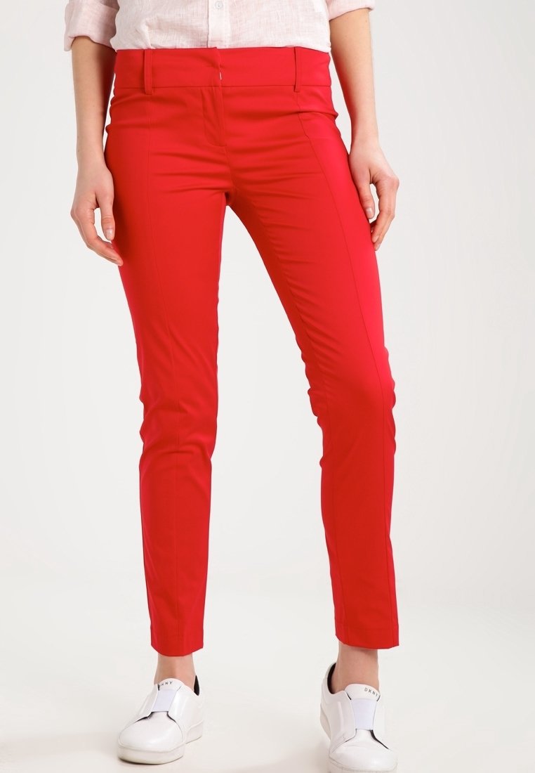 Femme portant un pantalon slim rouge vif et des baskets blanches sans lacets, debout devant un fond clair uni.
