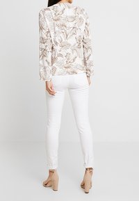 Blusa floreale a maniche lunghe in tessuto leggero, con motivi di foglie beige e verdi. Abbinata a jeans skinny bianchi e tacchi nudi.