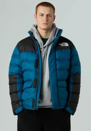 Giacca puffer blu e nera con zipper frontale, dettagli in Velcro e un piccolo logo sul petto, indossata sopra un hoodie grigio.