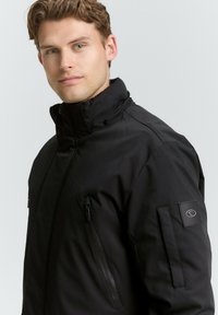 Schwarze, wasserdichte Jacke mit hohem Kragen, Reißverschlusstaschen, glatter Textur und einer Logo-Patch am Ärmel. Entworfen für Wärme und Schutz.