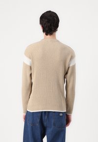 Beiger gerippter Pullover mit langen Ärmeln und weißen Streifenakzenten an den Schultern, mit einem runden Ausschnitt und strukturiertem Material.