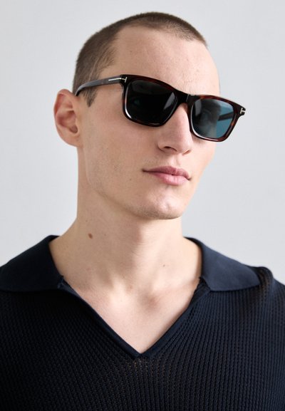 Jeune homme portant des lunettes de soleil écaille et un pull à col en V texturé foncé avec col, regardant légèrement sur le côté sur un fond uni.