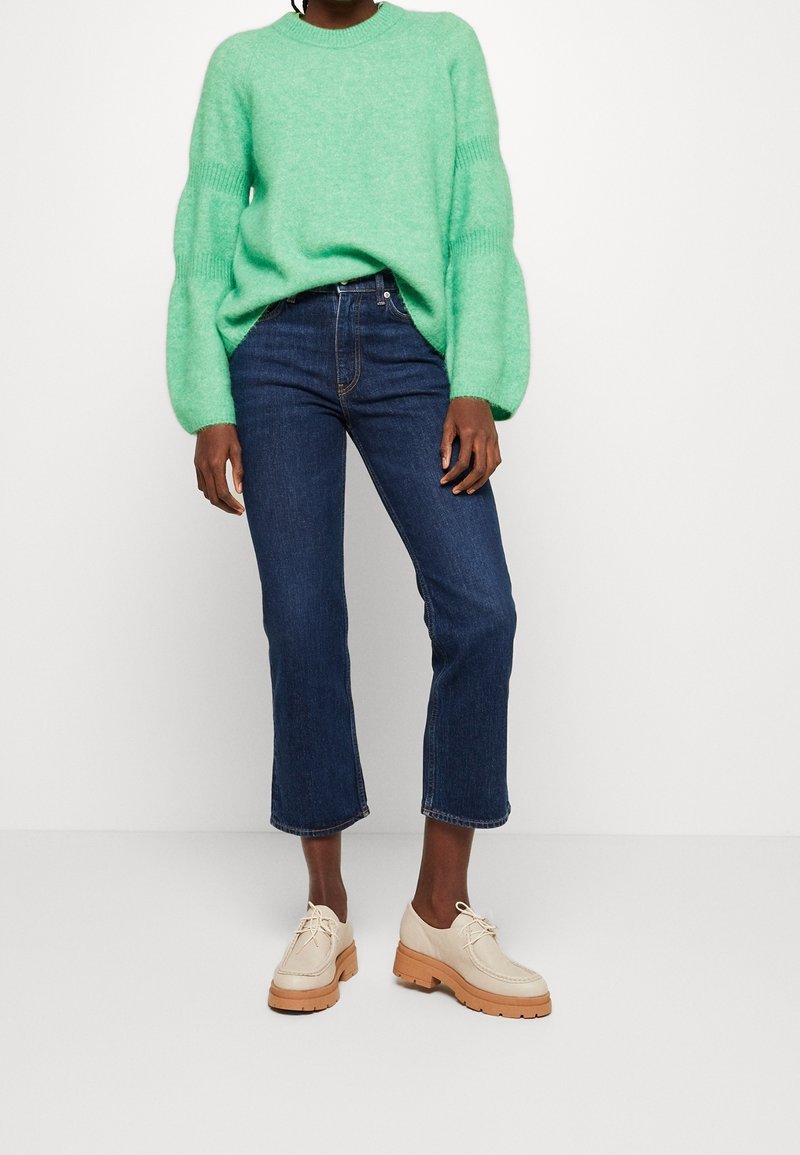 Personne portant un pull duveteux vert vif, un jean court bleu foncé et des chaussures beiges à semelles épaisses lacées, debout devant un fond blanc.