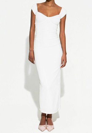 Femme portant une robe midi blanche ajustée épaules dénudées et des ballerines pointues roses claires avec des nœuds, debout devant un fond blanc uni.