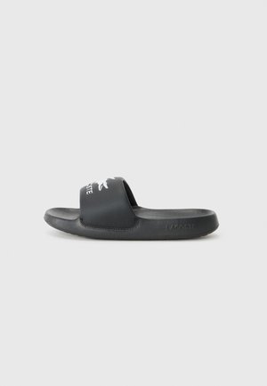 Black slide sandal with a wide strap featuring a white logo print. Textured footbed and smooth sole. Made of synthetic material.