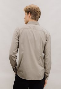 Lys grå langermet skjorte med krage, teksturert stoff og folder bak. Har knappede mansjetter og en enkel logo over kragen.