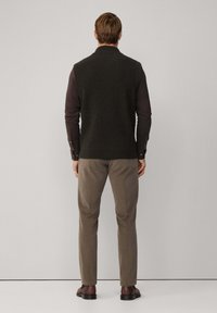 Tmavě zelená pletená vesta přes dlouhý rukáv hnědého trička, zkombinovaná se světle šedými kalhotami a tmavě hnědými botami. Jednoduchý design a přiléhavý střih.