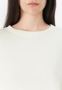 Light cream-colored top featuring a round neckline and smooth texture, with subtle stitching detail along the neck.